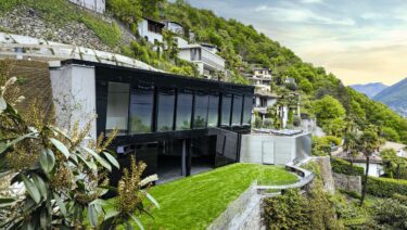 Das Ferienhaus fügt sich als nahezu schwarzer Kristallkörper in die Hanglage ein. Die dunklen, teils spiegelnden Glasflächen reflektieren Licht, Vegetation und Topografie.